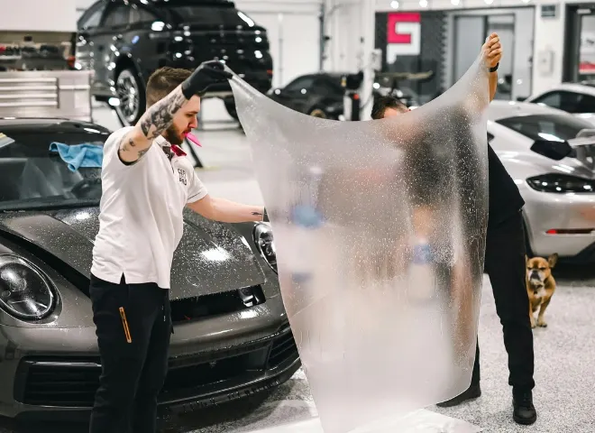 Two technicians install a clear protective film on a gray sports car's windshield inside a bright auto detailing shop.