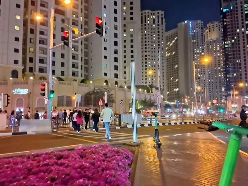 Nighttime urban street scene with a group of pedestrians waiting to cross a wide crosswalk, tall illuminated buildings in the background, and colorful street lights.